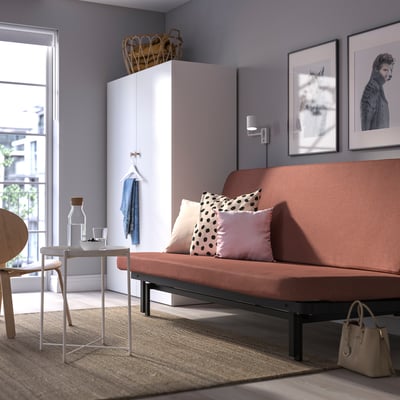 Modern living room with NYHAMN sofa-bed in terracotta, under a tall wardrobe, lit by a large window.