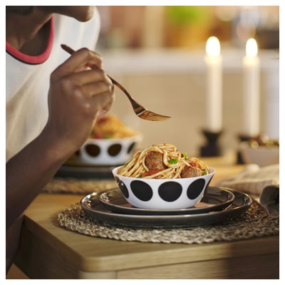 A person eats spaghetti from an OFTAST white bowl with black polka dots on a wooden table.