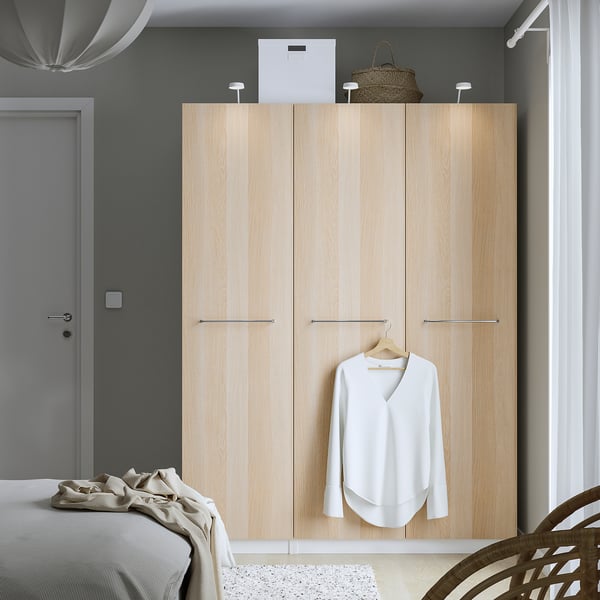 Minimalist bedroom with large light wooden PAX closet, white blouse hanging, woven bag atop, beside beige bed.