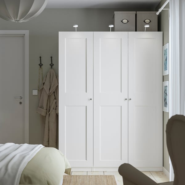 Minimalist bedroom with large white PAX wardrobe, silver handles, two grey boxes, narrow door, bed with white and beige linen.