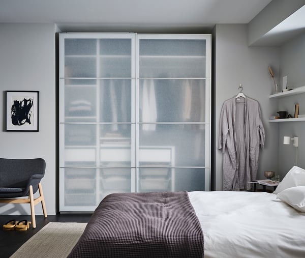 Modern bedroom with large PAX wardrobe, frosted glass doors, grey walls, white bed with grey blanket, robes hanging, grey rug, and abstract art.