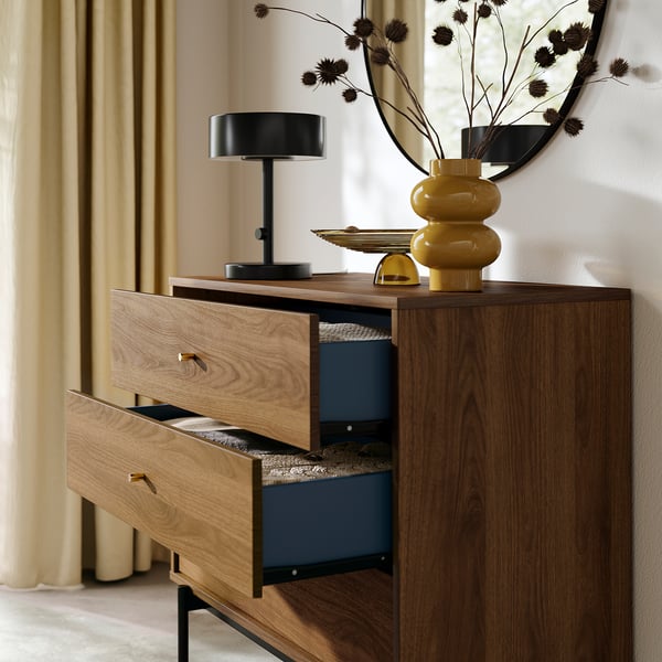 Wooden dresser with three open drawers, displaying interior, alongside black lamp and yellow vase on top, against wall with mirror.