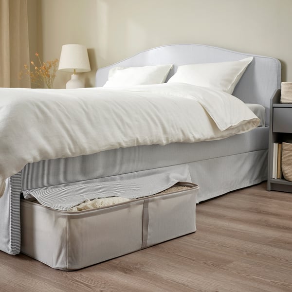 A neatly made bed with a curved headboard, white bedding, and striped bed skirt showing under-bed storage with a visible box.