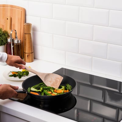 Person cooking vegetables in a frying pan with a spatula. Kitchen items on the countertop, including a cutting board.