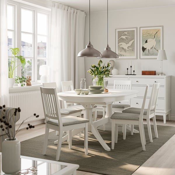 White extendable dining table with 6 grey chairs, 2 pendant lights, in bright room.