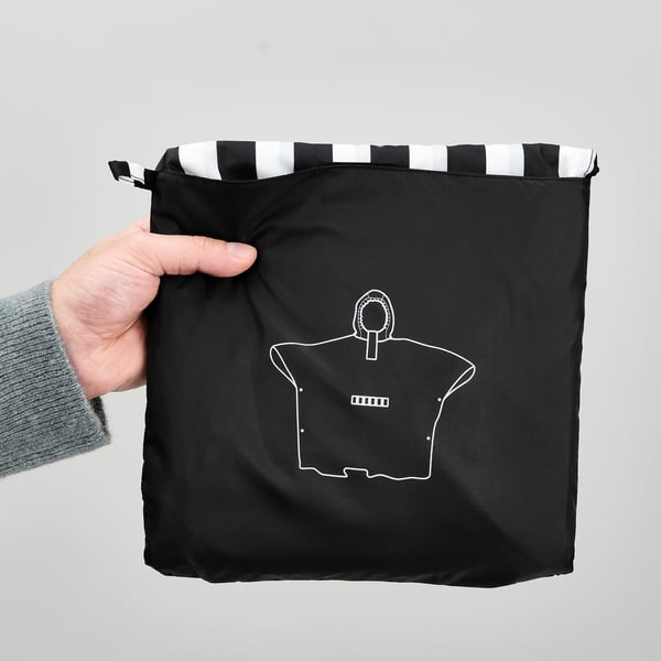 Hand holding black poncho with white stripes at top, featuring a white outline of a shirt with a pocket.