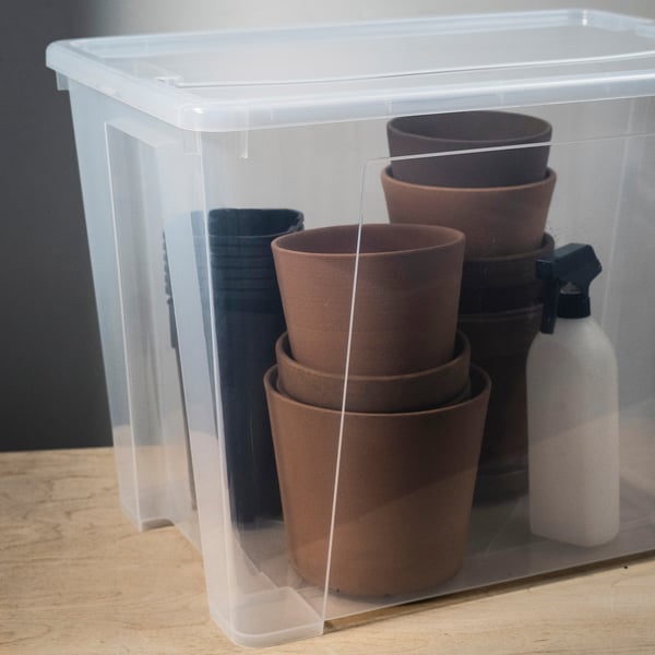 Clear plastic storage box with snap-lock lid, holding stacks of terracotta pots and a spray bottle, neatly organised.