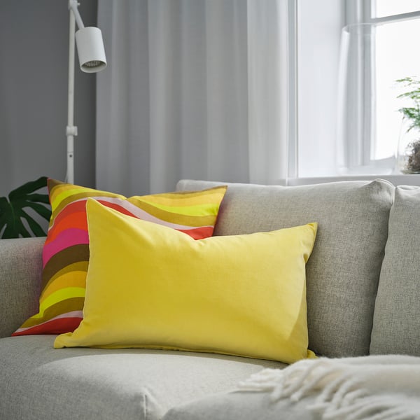 Yellow velvet cushion on sofa, colourful pillow behind, grey curtain, lamp, plant.