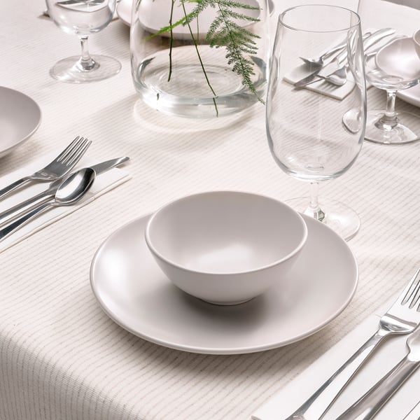 A dining table set with white plates, bowls, silverware and glasses. A vase with greenery is centred. A beige, textured tablecloth covers the table.