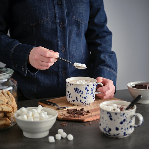 Person adding marshmallows to mug from bowl on dark surface.