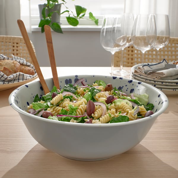 A pasta salad in a white bowl on a table, with wooden tongs, glasses, and bread basket nearby.