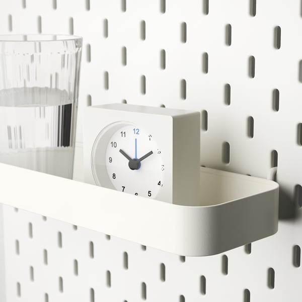 White shelf with clock and container attached to pegboard. Holds small items.