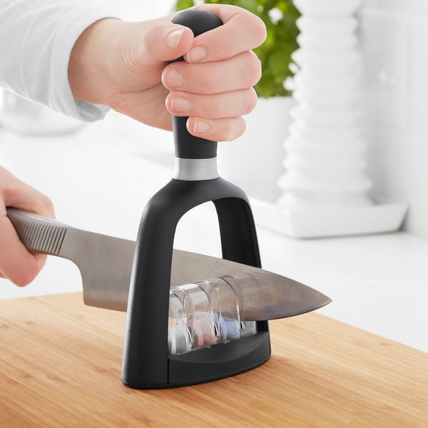 A person uses a black SKÄRANDE sharpener to sharpen a knife on wood. Multiple slots for different sharpening levels.