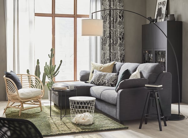 Modern living room with grey SKAFTET floor lamp, cushioned grey sofa, wicker chair, green rug, and large window with black patterned curtains.