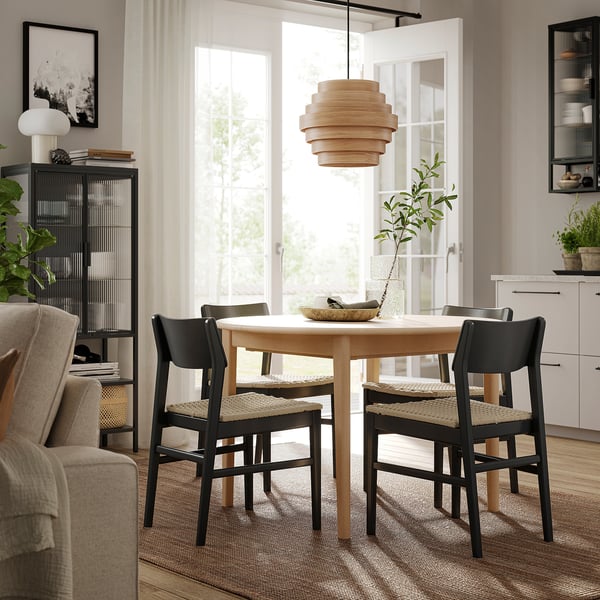 Modern dining area with round light wood table, hanging light fixture, dark chairs, large window, beige rug, and black bookshelf.