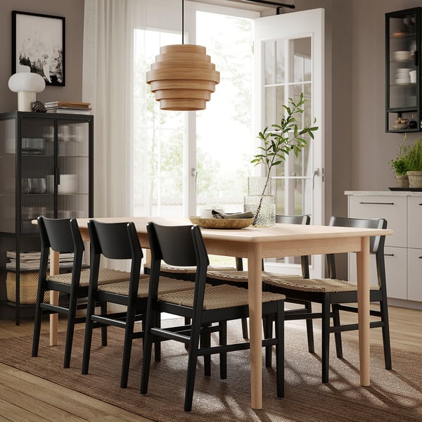 Modern dining area with light wood table, four black chairs with woven seats, hanging wooden lamp, and large window.