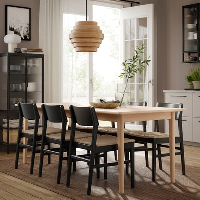 Modern dining area with light wood table, four black chairs with woven seats, hanging wooden lamp, and large window.