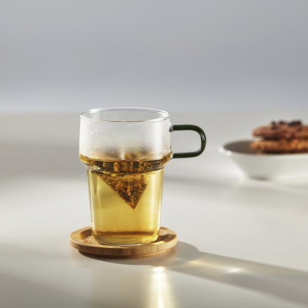 Clear glass mug with green handle holds tea bag, sits on wooden coaster on white table.