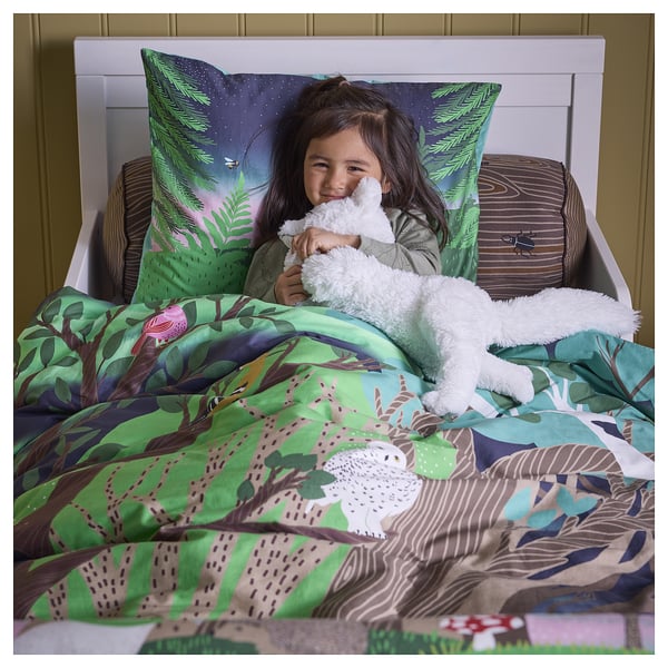 A child hugs a soft white fox toy in bed, covered by a green forest-themed blanket.