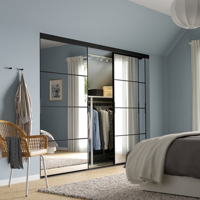 Modern bedroom with large black SKYTTA closet, mirrored panels, grey & white bed, wicker chair, and patterned rug.
