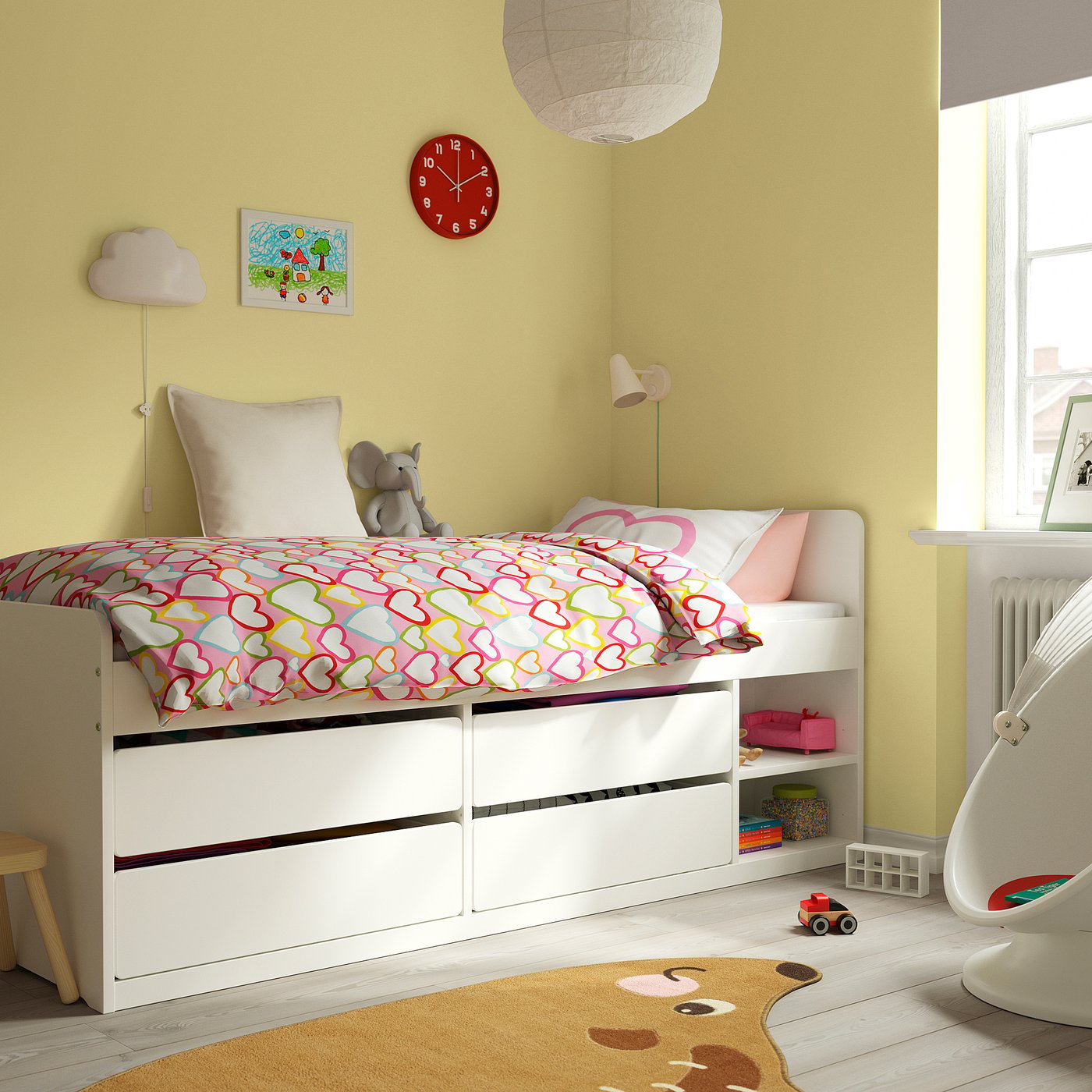 Kids room with yellow walls, white bed, heart bedding, toy, clock, lamp, rug, bookshelf.