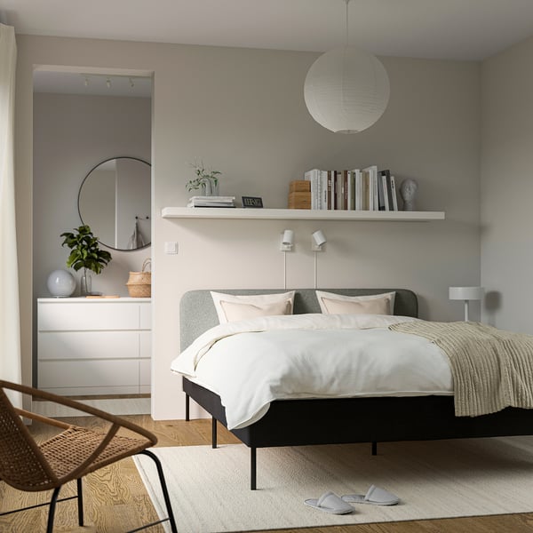 Modern bedroom with dark grey SLATTUM bed, soft headboard, bookshelf, dresser, mirror, wicker chair, beige rug.