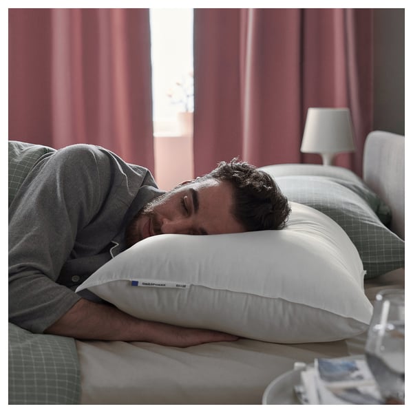 A person sleeps peacefully on a white SMÅSPORRE pillow with a grey and white chequered pillowcase in a cosy bedroom with red curtains.