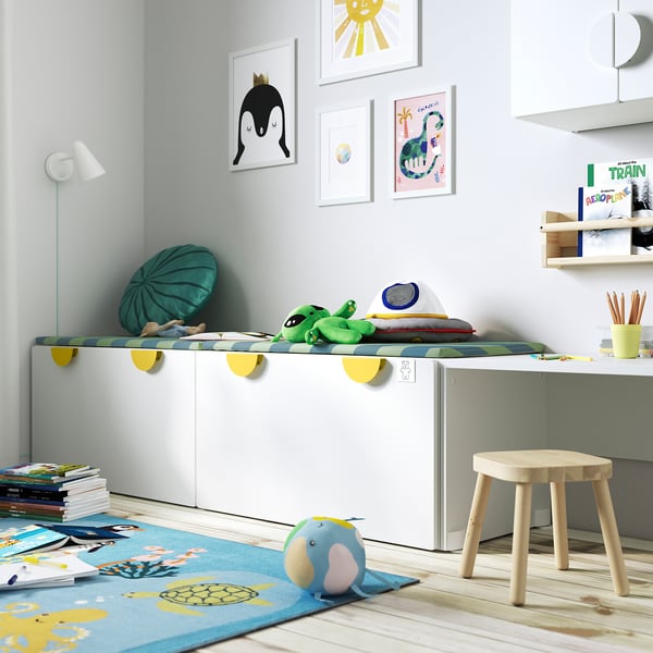 Kids playroom with SMÅSTAD toy storage bench, green cushion, colourful art, blue rug, wooden furniture.