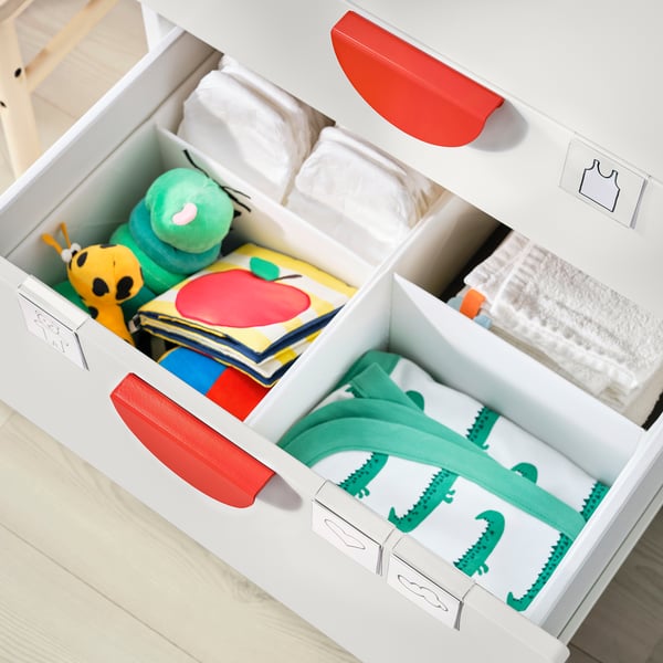 White nursery organiser with red handles, storing baby clothes and toys, including green dinosaur pyjamas.
