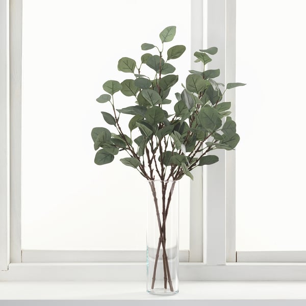 Glass vase on window sill with artificial eucalyptus. Alternative: vase with artificial eucalyptus on window sill.