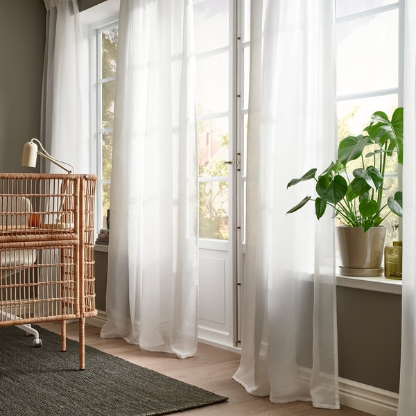 Bright living room with STENFRÖ sheer white curtains. Large wooden door, wicker bookshelf, plant on table. Clean, minimalist design.