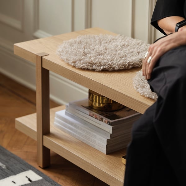 Wooden side table with natural finish supports persons arm, stacked books and a cosy cushion on shelves.