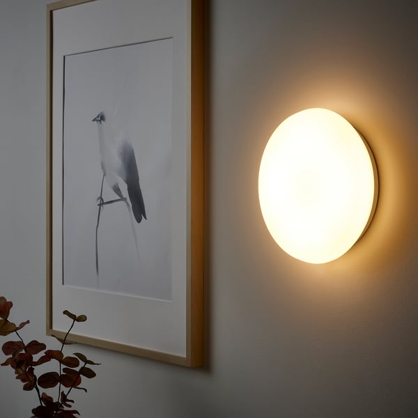 A round, wall-mounted light fixture emitting warm-white light, part of IKEAs smart lighting system.