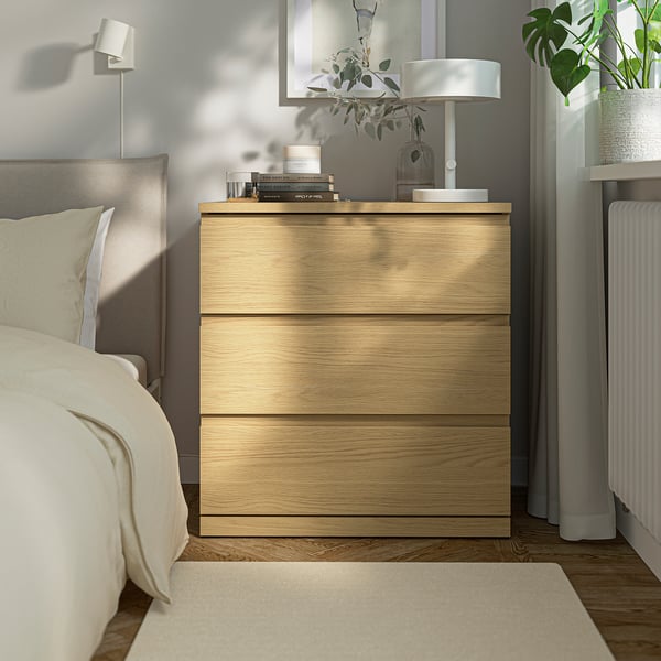 Oak chest of drawers in bedroom, three drawers, beside bed with lamp & plants.