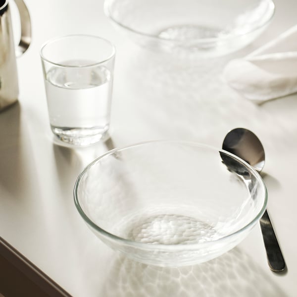 STRANDKRABBA bowl, clear glass, small & round, hammered texture, shiny spoon, water glass & blurred background.