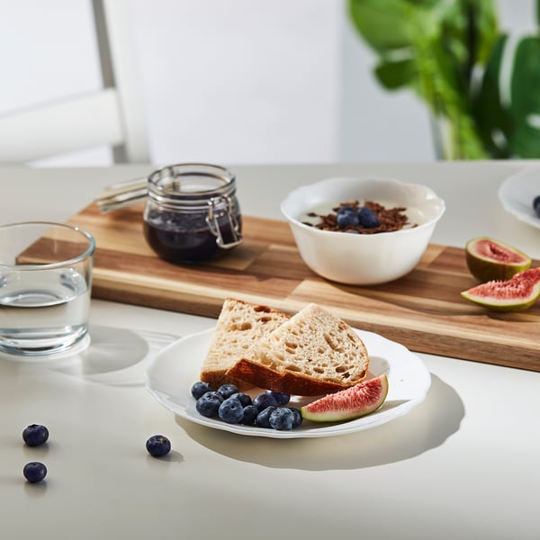 White plate with toast and fruits on a wooden board beside a glass jar and a bowl with yoghurt and granola, all on a white countertop.