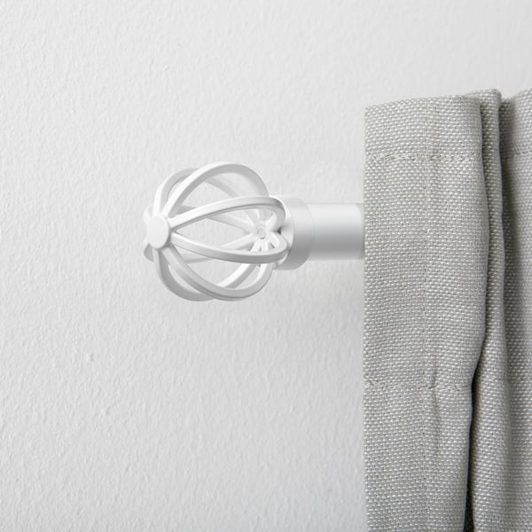A white, circular, metal curtain rod finial. Its small, positioned next to a larger, light grey curtain.
