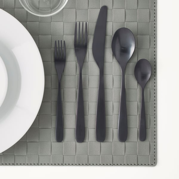 Modern black cutlery set on grey place mat, including fork, knife, and spoon, shown with white plate and glass.