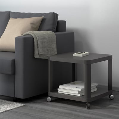 Dark grey sofa with light-eye cushion, grey throw over arm. Beside it, black rolling side table with books and remote on top.