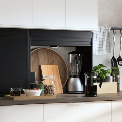 Modern kitchen with TITTEBO hidden storage. Black tambour door, metal appliances, wood cutting boards, and plants.