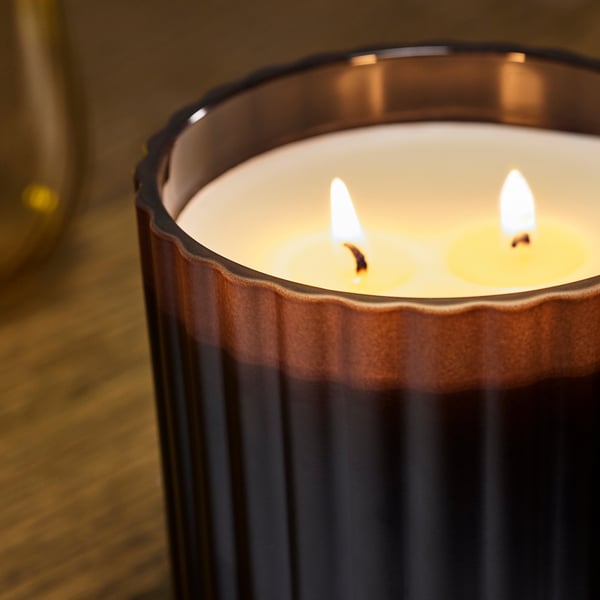 Three wicks of a scented candle burn steadily in a ribbed glass container, emitting a warm and spicy aroma.