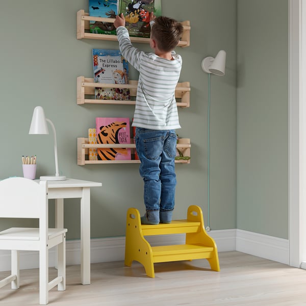Child standing on yellow TROGEN stool, reaching for books on wall shelves. White desk and lamp nearby.