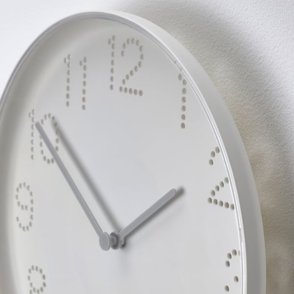White round wall clock with punched hole numbers, hands pointing at twelve and six. Minimalist design, mounted on a plain wall.