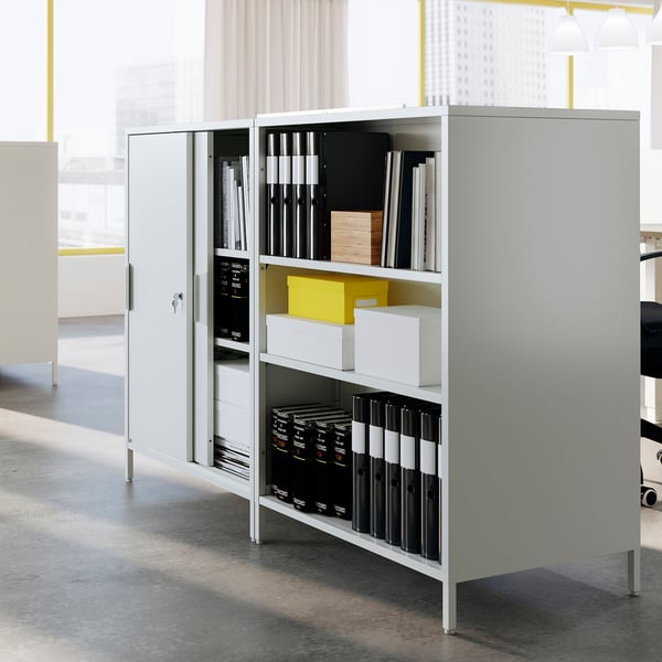A white TROTTEN double-sided office cabinet with sliding doors, storing binders and boxes, acting as a room divider.