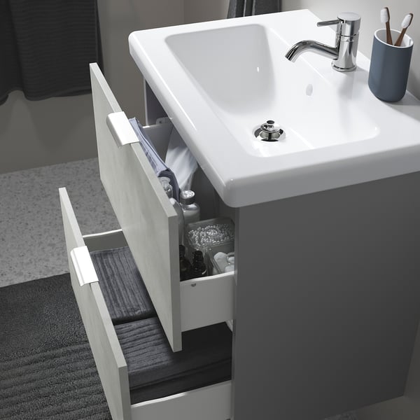 Modern bathroom sink with open drawer showing toothbrush holder and toiletries. Tap and grey countertop visible.