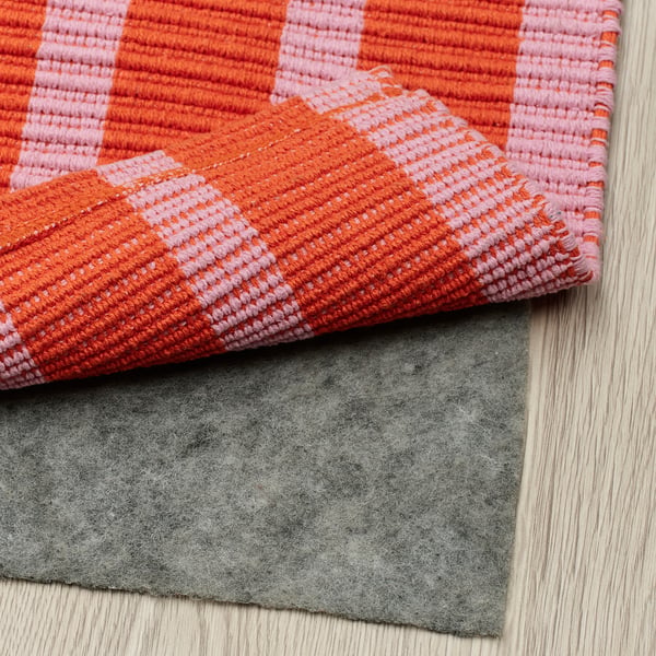 A rug with pink and orange stripes is laid over a grey anti-slip mat on a wooden floor.