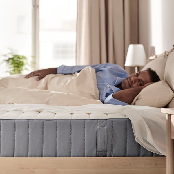 A person sleeping on a blue and white VALEVÅG mattress. The room is peaceful, with neutral curtains and a lamp on a wooden bedside table.