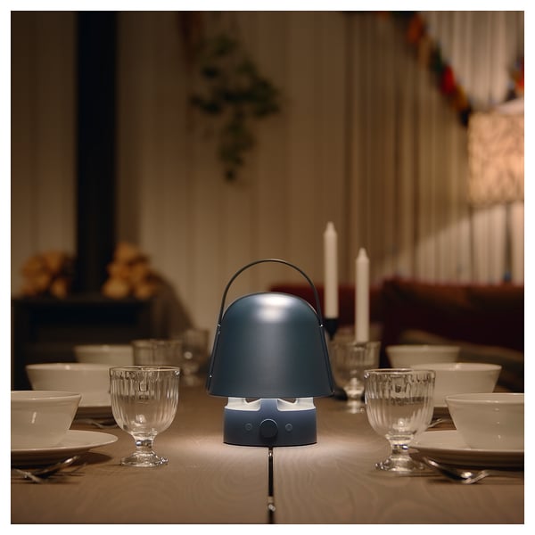 A blue VAPPEBY speaker lamp sits on a dinner table. It has a handle and emits a warm light. Surrounded by glasses, plates, and candles.