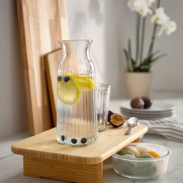 Carafe of lemonade with berries and lemon slices on wooden board, with glass, plates, fruits, and plant in background.