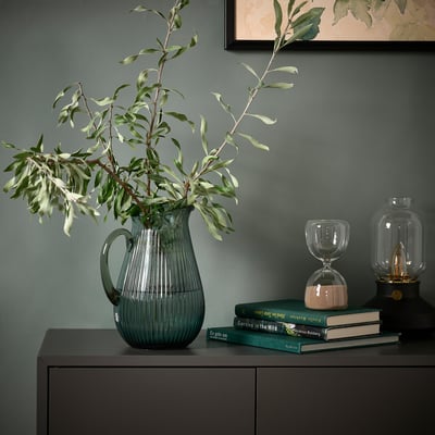VARDAGEN green glass jug on dark cabinet with branches, books, hourglass.